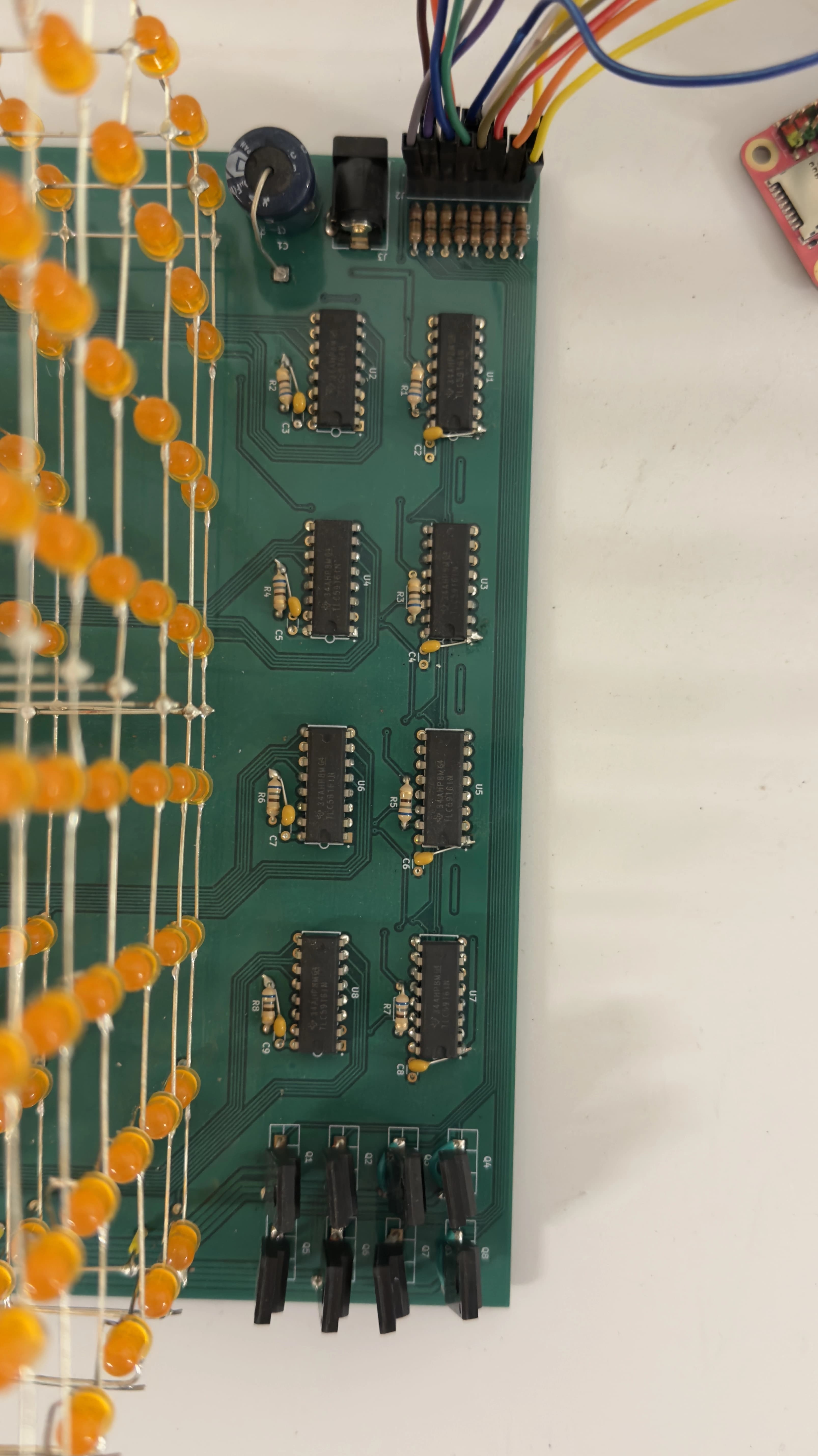 Assembled PCB beside the cube, populated with eight LED driver ICs, resistors, decoupling capacitors, pMOS transistors, a barrel jack, and a GPIO header with rainbow jumper wires