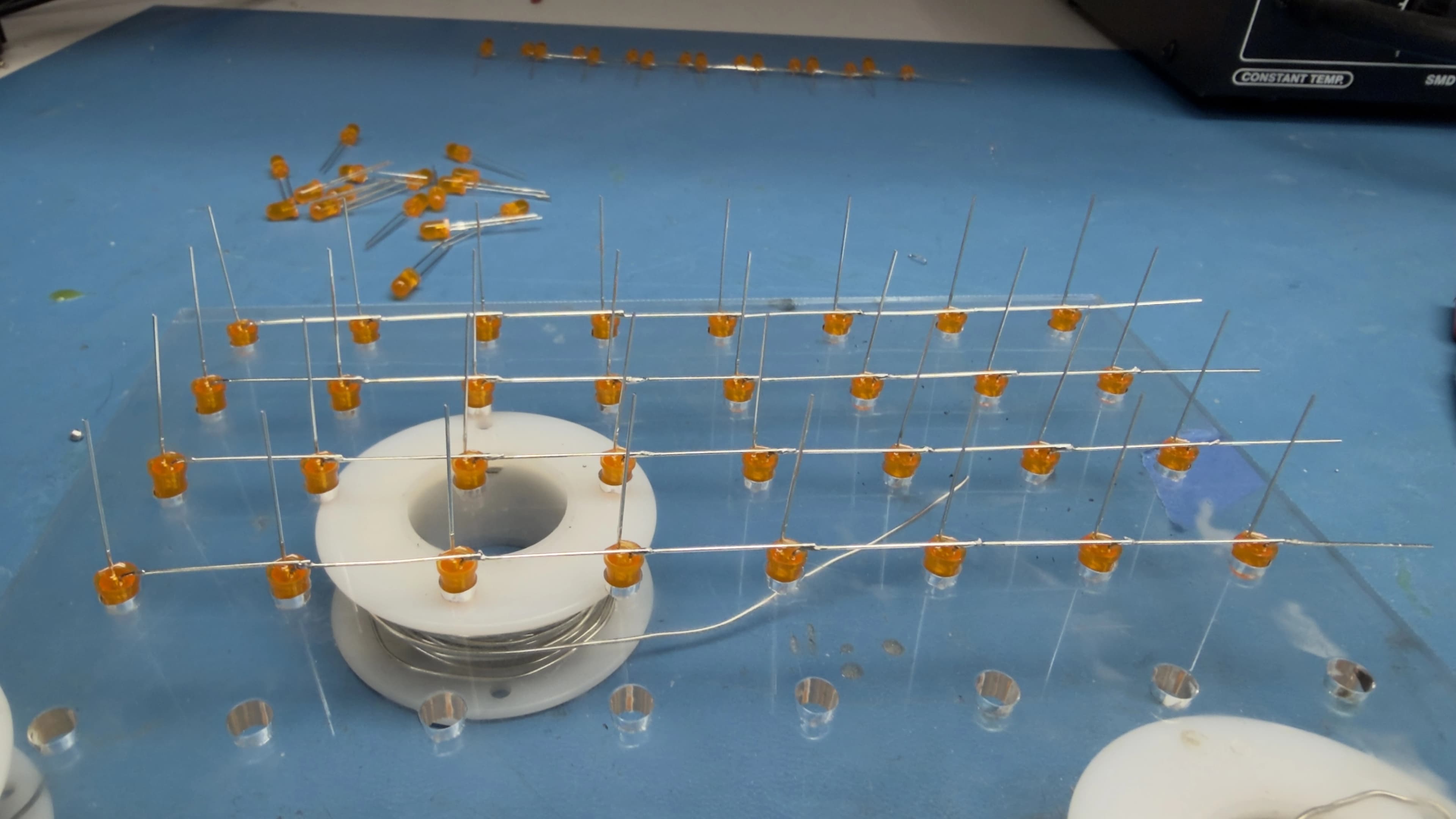 Orange LEDs seated in the acrylic jig with their bent anode leads soldered into rows, next to a spool of wire on a blue soldering mat