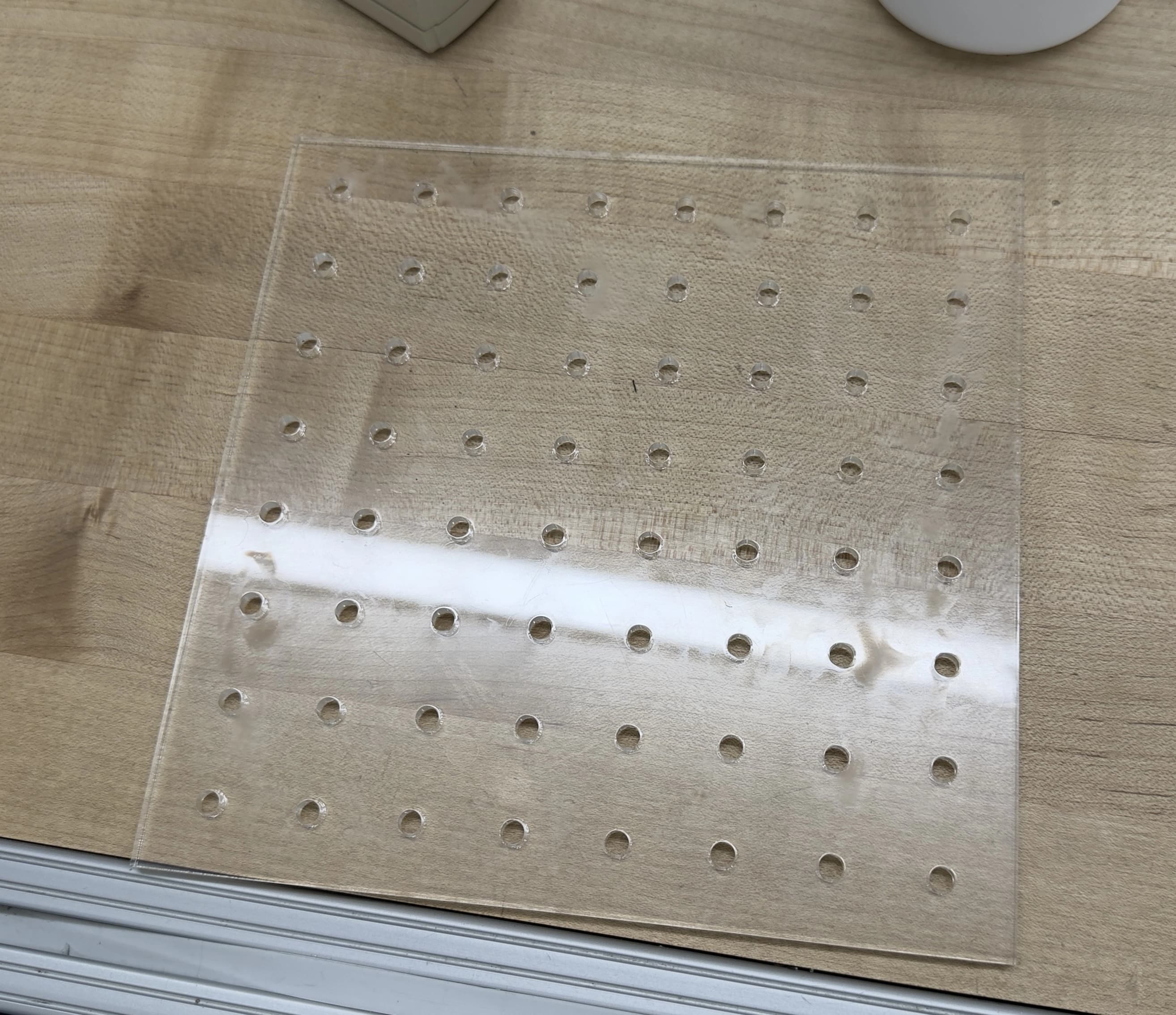 Clear laser-cut acrylic sheet with an 8x8 grid of holes sized to hold LEDs, lying on a wooden workbench