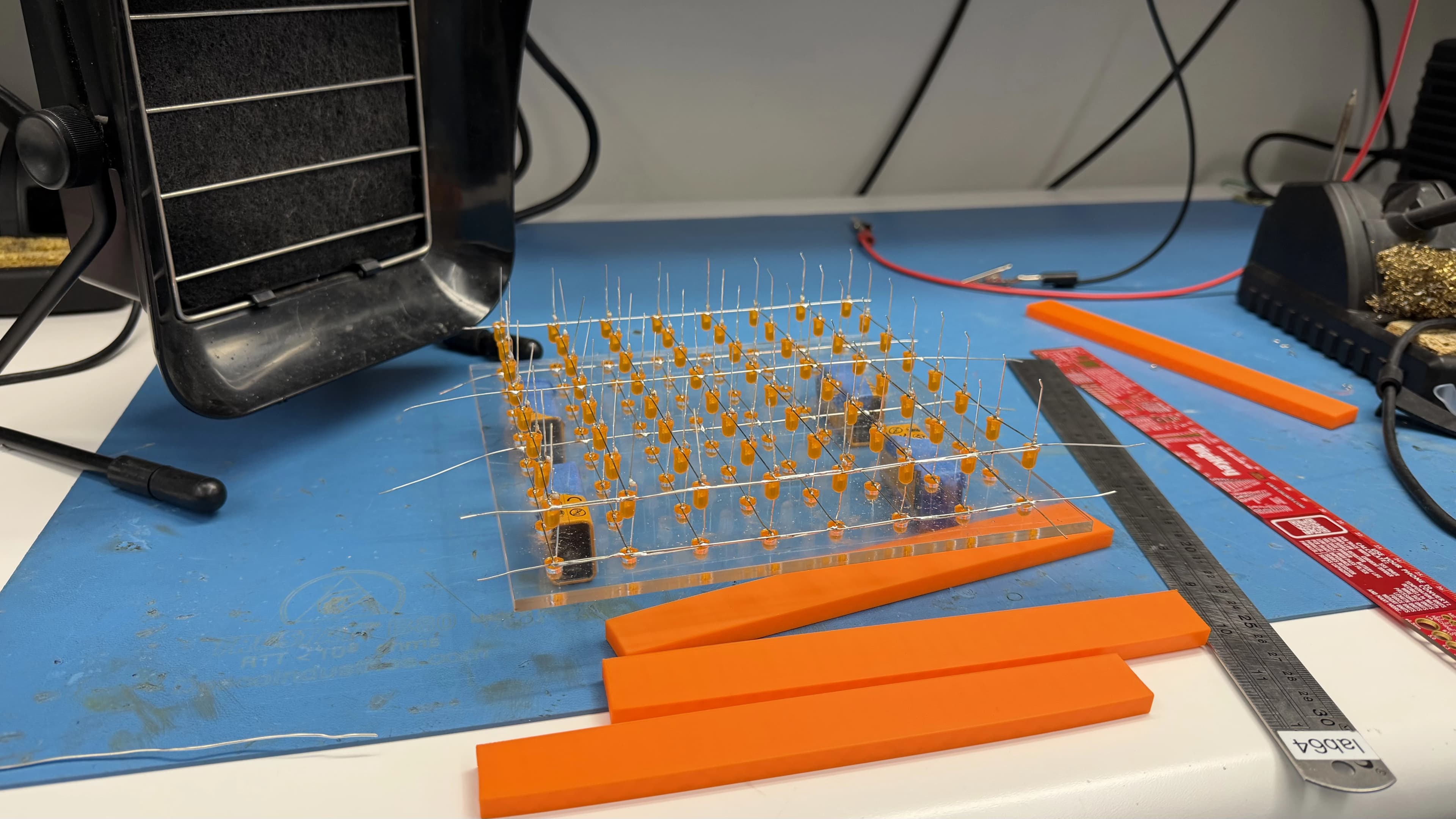 Orange 3D-printed spacer bars and a steel ruler beside an LED layer with upright cathode leads, sitting on the jig at a soldering station