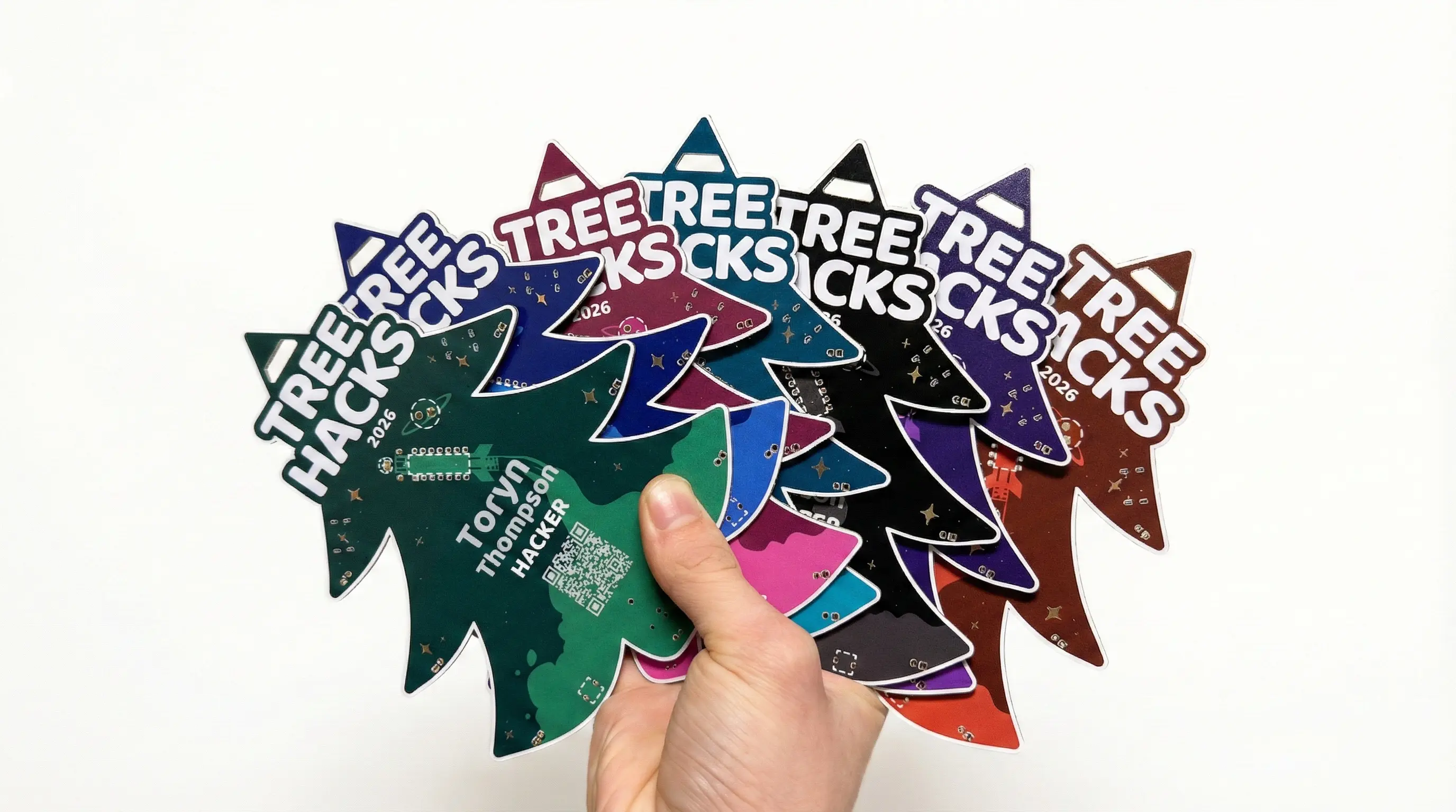 A hand fanning out tree-shaped TreeHacks 2026 PCB badges in several colors