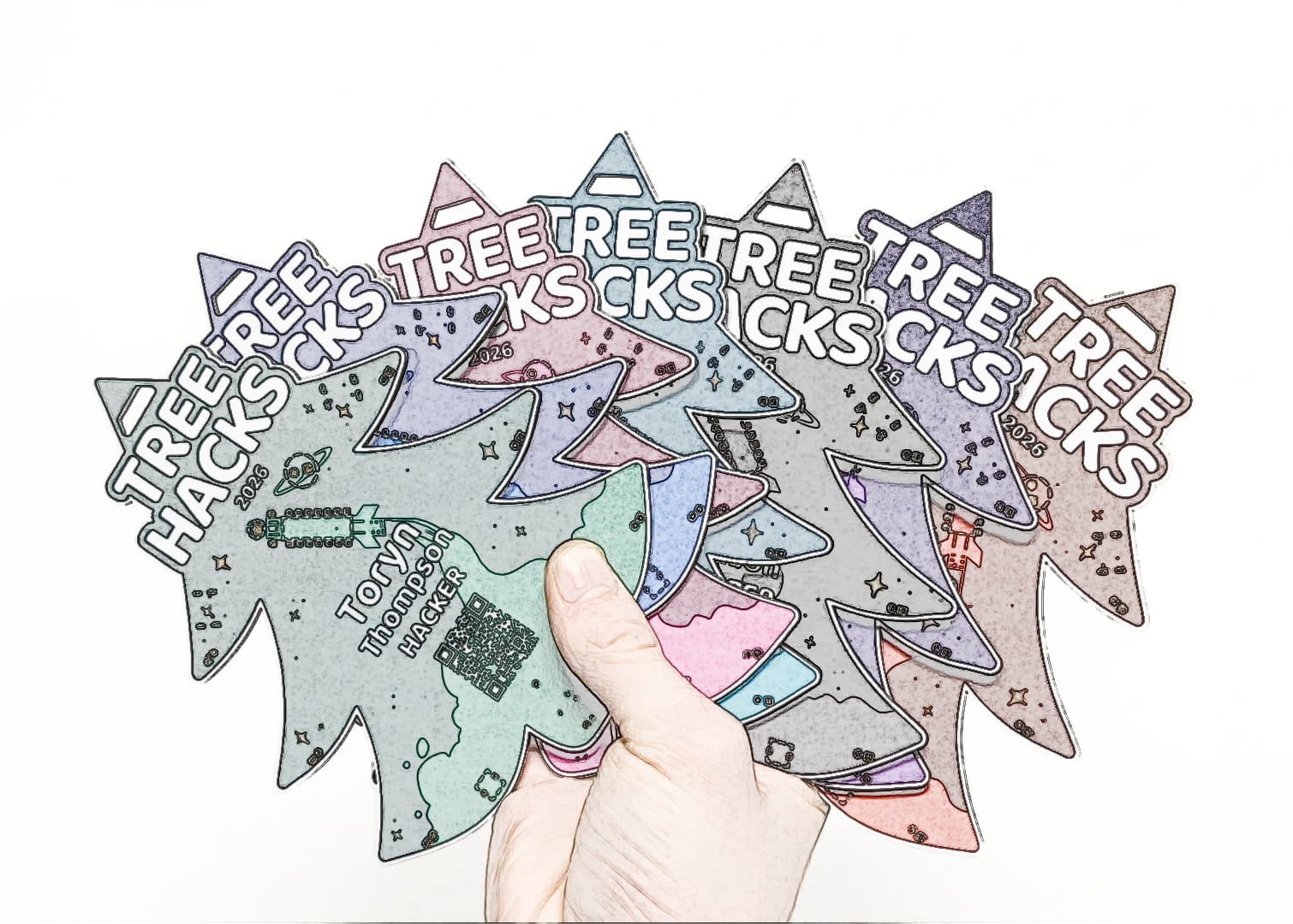 A hand fanning out tree-shaped TreeHacks 2026 PCB badges in several colors
